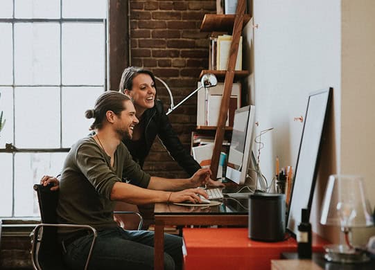 Two people are working together at a desk, looking at a computer screen and smiling in a modern office with exposed brick walls.