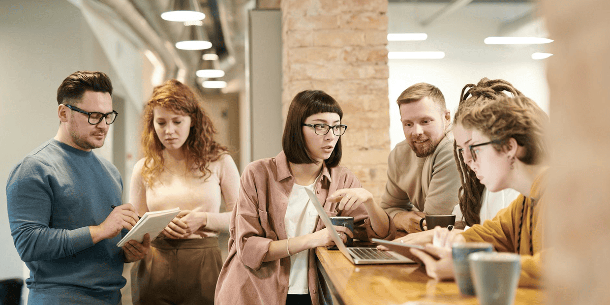 Five people gather around a wooden counter in an office, discussing and looking at a laptop and notebooks, with coffee cups nearby.