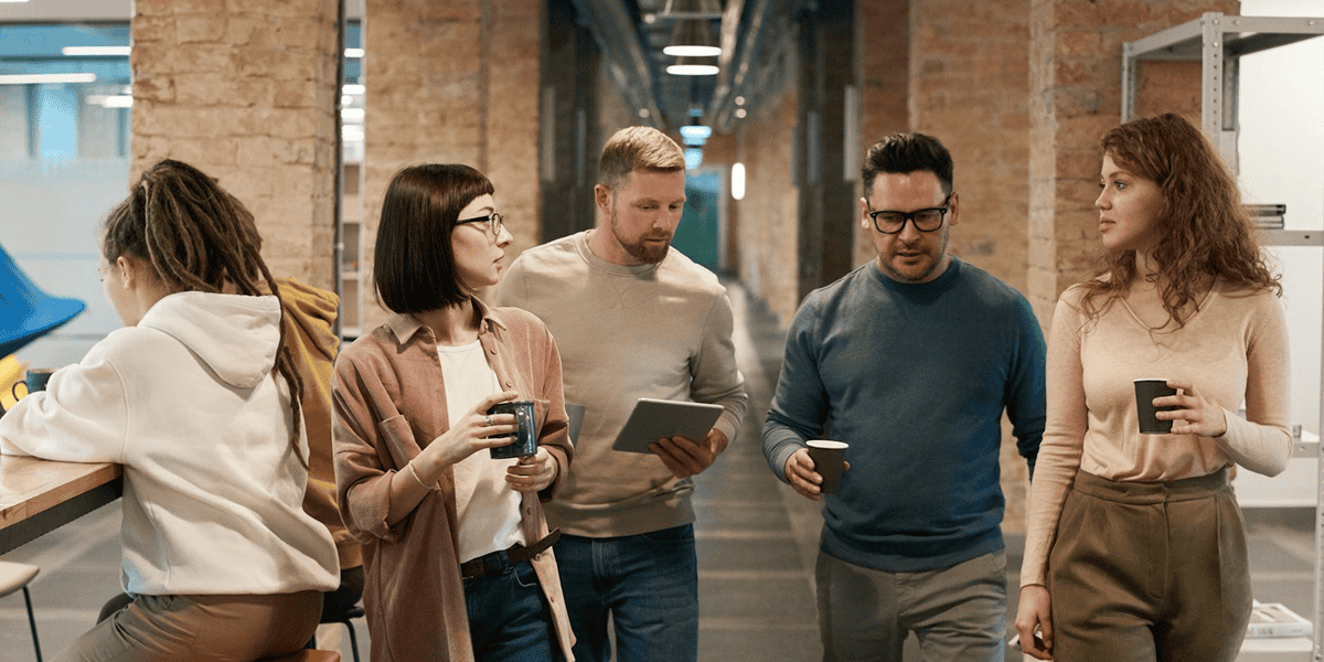Five people in casual clothing walk and talk in an office hallway; one uses a tablet, others hold cups, and one person sits at a counter with a laptop.