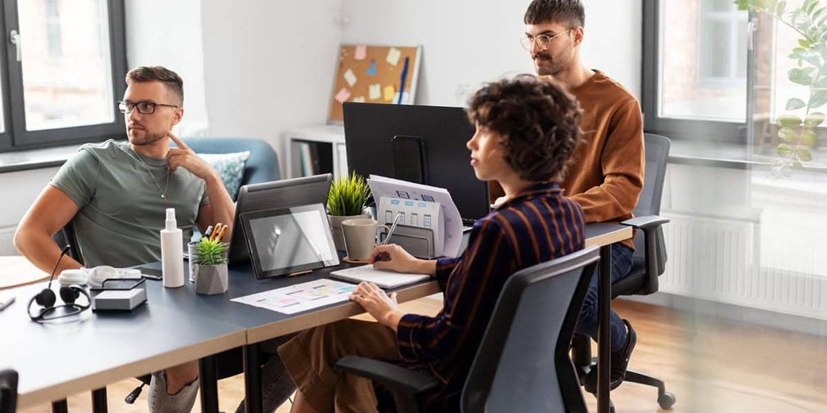Three people sit at a desk in a modern office, working with laptops, tablets, and documents, with plants and office supplies on the table.