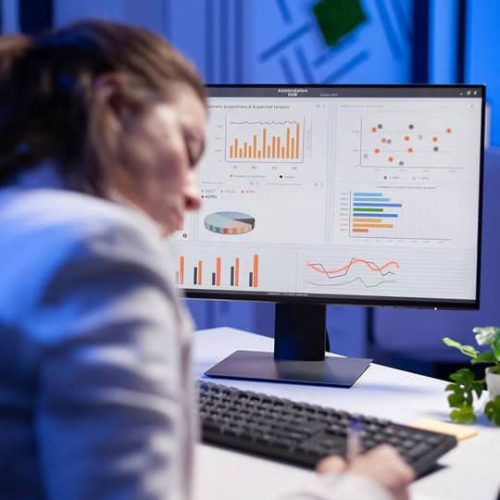 Person sitting at a desk, writing on paper, with a computer monitor displaying various colorful charts and graphs.
