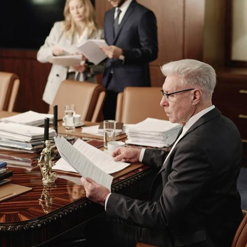 An older man in a suit reads documents at a conference table, while two younger colleagues review papers in the background.