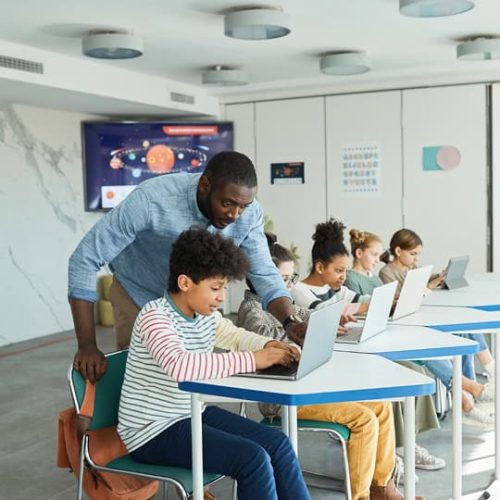 A teacher assists a student working on a laptop in a classroom, while other students also use laptops at their desks.