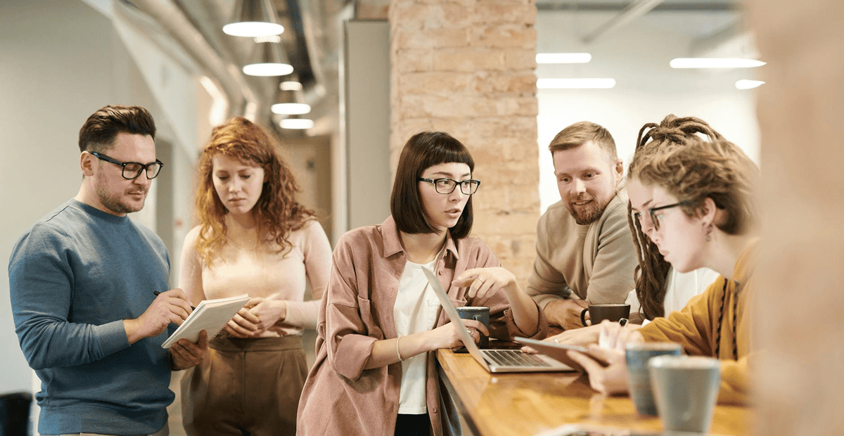 Five people gather around a wooden counter in an office, discussing and looking at a laptop and notebooks, with coffee cups nearby.