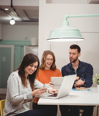 Three people sit at a table working with a laptop and tablet, appearing to discuss or collaborate in a modern office setting.
