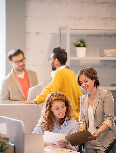 Four people work in a modern office; two women review documents at a desk in the foreground, while two men talk in the background.