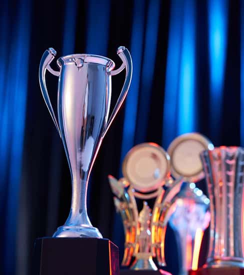A close-up of a silver trophy cup in focus, with several other gold and crystal trophies blurred in the background against a blue curtain.