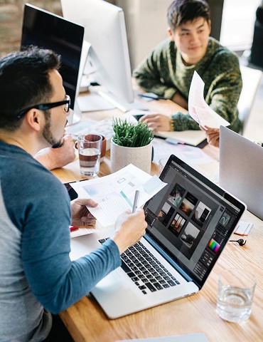 Two people sit at a desk with laptops and documents, discussing work in a modern office setting. A plant and glasses of water are on the table.