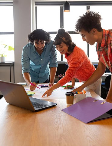 Three people stand around a table in an office, looking at papers and a laptop, discussing work. A coffee cup and purple folder are also on the table.