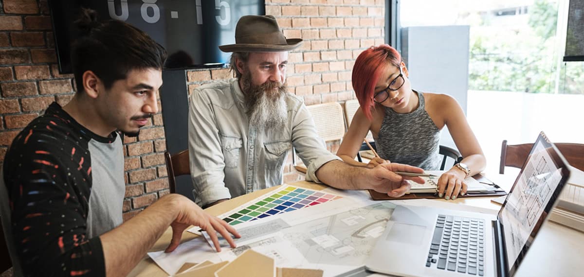 Three people sit at a table discussing design samples and color swatches while looking at a laptop in a modern office with a brick wall.