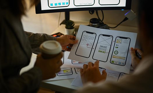 Two people review printed mobile app wireframes at a desk, one holding a coffee cup, with a computer monitor and plant in the background.
