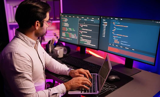 A man sits at a desk working on a laptop, with two large monitors displaying code in a dimly lit office with purple and pink lighting.