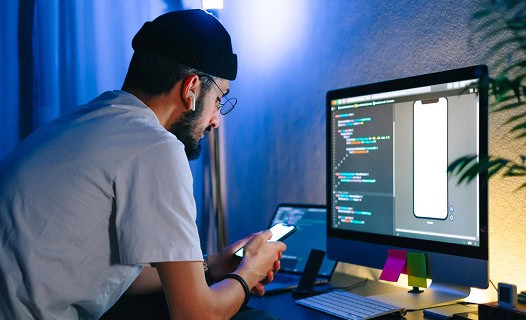 A person wearing glasses and a beanie sits at a desk, looking at a smartphone while computer code and a smartphone interface appear on a monitor.