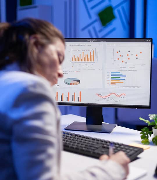 Person sitting at a desk, writing on paper, with a computer monitor displaying various colorful charts and graphs.