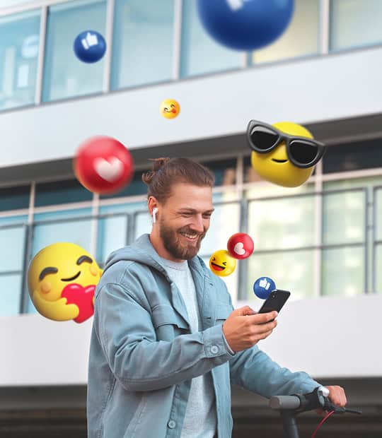 A man in a light jacket looks at his phone and smiles, surrounded by floating social media and emoji icons against a modern building background.
