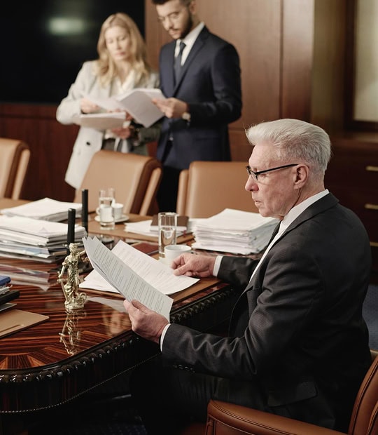 An older man in a suit reads documents at a conference table, while two younger colleagues review papers in the background.