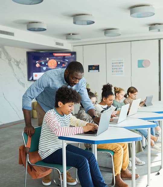 A teacher assists a student working on a laptop in a classroom, while other students also use laptops at their desks.