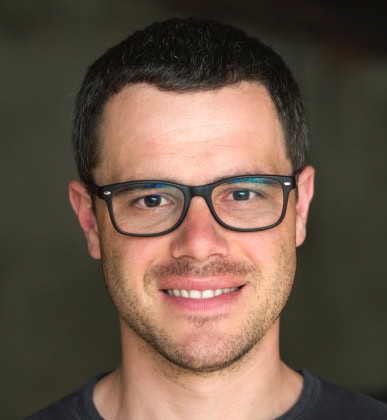 A man with short dark hair, glasses, and slight facial hair smiles at the camera against a blurred, dark background.