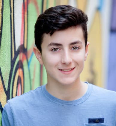 A teenage boy with short dark hair wearing a light blue shirt stands in front of a colorful graffiti wall, smiling at the camera.