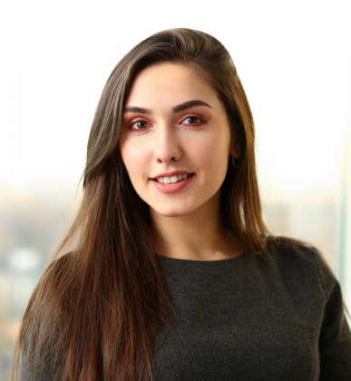 A young woman with long brown hair, wearing a dark gray top, smiles at the camera against a bright, blurred background.