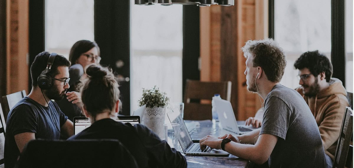 Five people sit around a table working on laptops in a well-lit room, appearing focused and engaged in discussion.