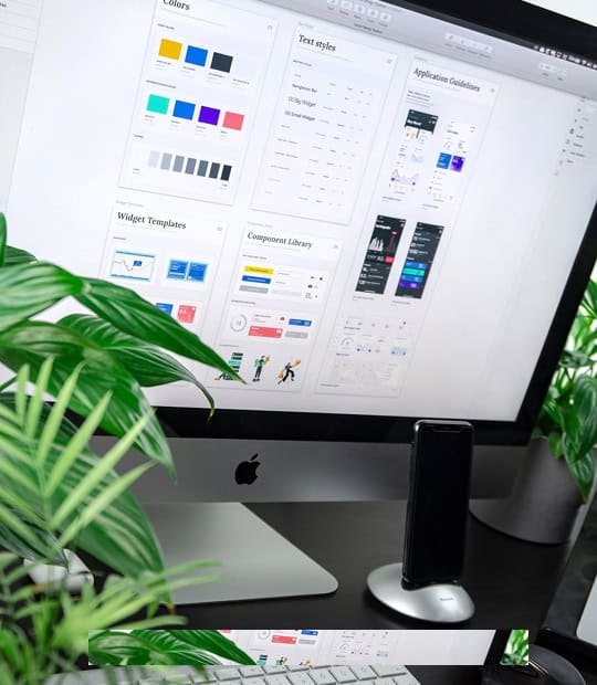 A desktop computer displays a design system interface with color palettes, text styles, and app guidelines; plants and a phone are on the desk.